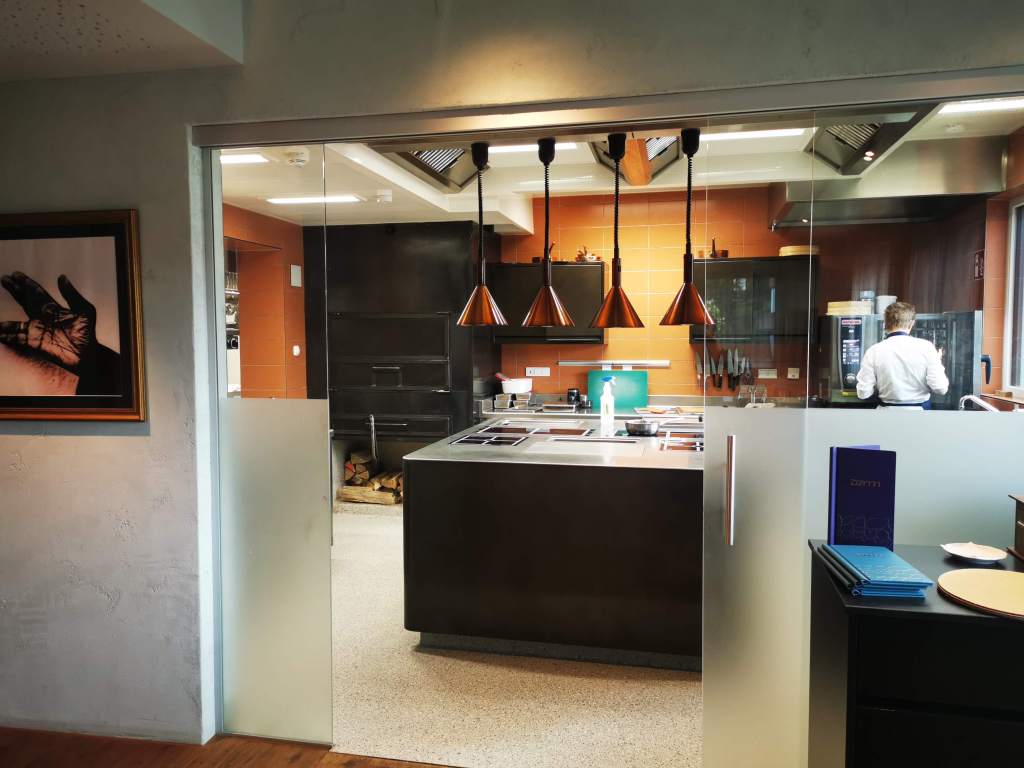 Modern kitchen interior with a large central island, stainless steel appliances, and stylish pendant lights. A chef in a white jacket is preparing food in the background.